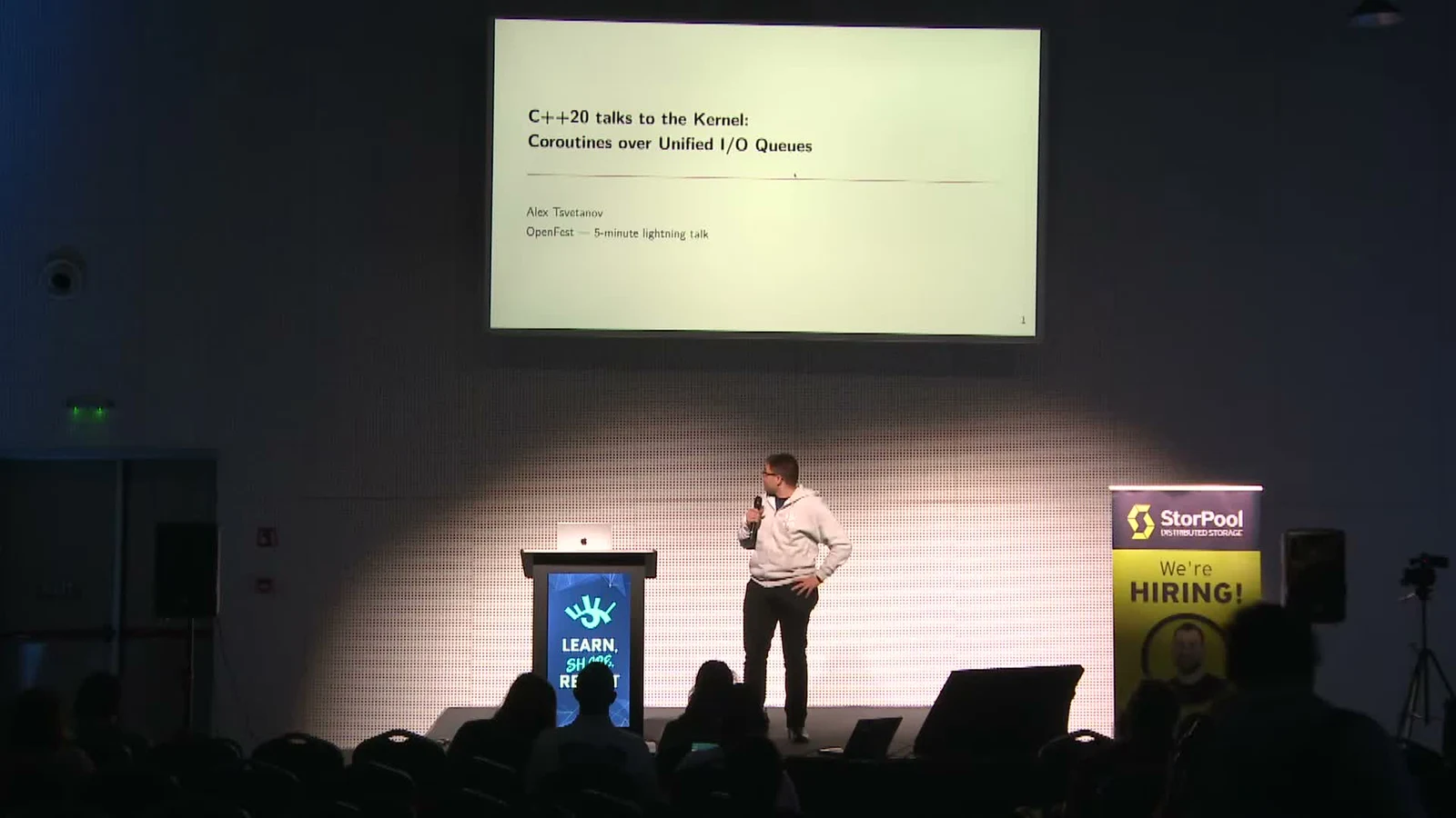 Alex presenting at OpenFest 2025, with the stage and audience in the foreground